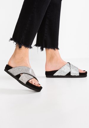 Fødder iført sorte åbne slide-sandaler med glitrende sølv-krydsseler, kombineret med sorte bukser med flossede kanter.