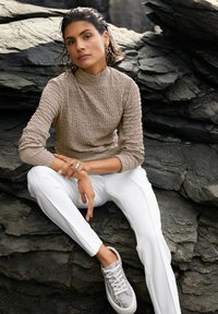 Frau mit nassen, kurzen Haaren, die auf dunklen Felsen sitzt und ein beigefarbenes strukturiertes Langarmshirt, weiße Hosen, Sneakers mit Schlangen-Muster und Schlangen-förmige Ohrringe trägt.