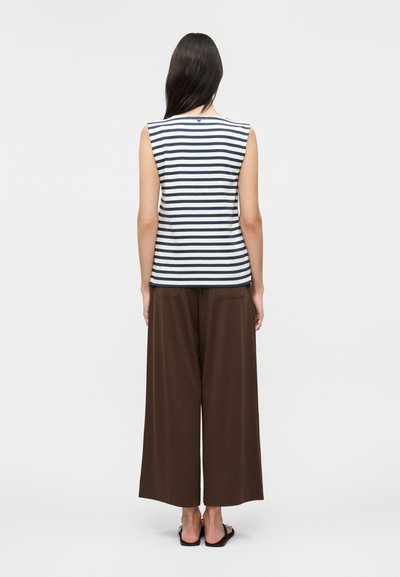 Femme aux longs cheveux foncés portant un haut sans manches blanc et bleu marine à rayures, un pantalon large marron et des sandales noires, debout de dos.