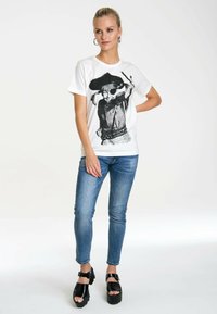 White cotton T-shirt featuring a black graphic print of a child wearing a hat, paired with blue skinny jeans and black platform sandals.