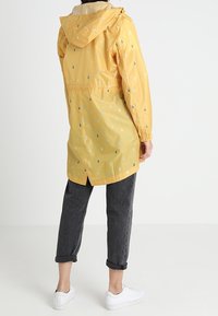 Gul regnrock med droppmönster, lätt material, justerbar huva, midjedragkedja och långa ärmar. Kombinerad med korta mörka byxor och vita skor.