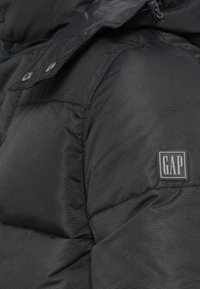 Melnā polsterētā jaka ar spīdošu tekstūru, ar apkakli, kas aizdara ar spiedpogām, un kvadrātveida GAP logo uzpatu plecā.