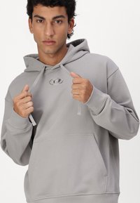 Hellgrauer Hoodie aus weichem Stoff mit einer Fronttasche, Kordeln und einem metallischen Logo auf der Brust. Mit gerippten Bündchen.