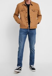 Béžová džínová bunda se zapínáním na knoflíky a dvěma náprsními kapsami, v kombinaci s modrými džíny a černobílými teniskami.