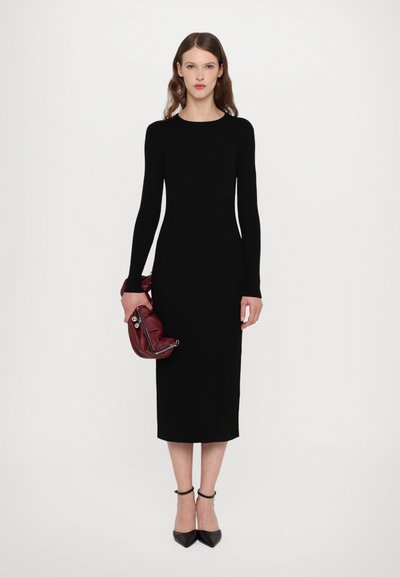 Vestido negro de manga larga con textura acanalada y silueta ajustada. La modelo sostiene un bolso burdeos y lleva puestos unos tacones de punta negra. Diseño sencillo y elegante.