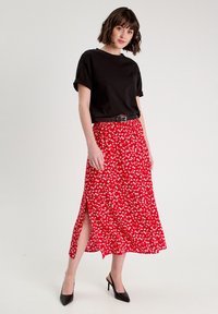 T-shirt en coton noir avec des manches roulées, associé à une jupe midi à imprimé floral rouge avec une fente sur le côté et une ceinture noire, complété par des talons noirs.