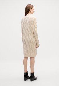 Beige kötött ruha magas gallérral, texturált mintával, combközépig ér, fekete bokacsizmával párosítva.