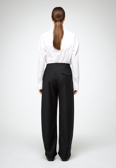 Vrouw met lang bruin haar in een paardenstaart, gekleed in een witte longsleeve en zwarte wijde broek, gezien van achteren, staande.