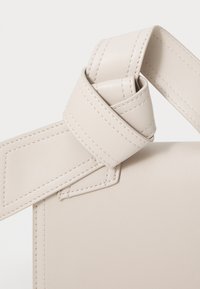 Gros plan sur un sac à main en cuir beige clair montrant une finition texturée, une bandoulière plate avec un détail plié et des coutures soignées le long des bords.