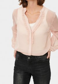 Helles rosa Bluse mit rüschigem Ausschnitt, transparenter Stoff und langen Ärmeln. Kombiniert mit dunkelblauen Jeans mit einer Vorderknopf.