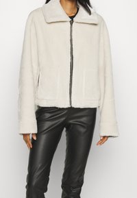 Modell i en ljusgräddfärgad kort jacka med krage, i kombination med svarta tighta byxor, stående mot en helt vit bakgrund.