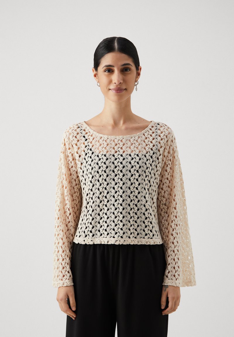 Haut en crochet avec un motif floral crème et noir, un décolleté bateau et de longues manches évasées, associé à un pantalon noir.