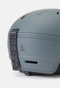Casco in-mold gris con una carcasa de policarbonato, ranuras de ventilación, hebilla ajustable e información de seguridad impresa en el costado.