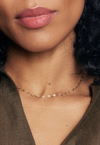 Złoty naszyjnik łańcuszkowy z naprzemiennymi ogniwami w kształcie owalu i okręgu, delikatnie spoczywający na gładkiej powierzchni skóry, z subtelnym połyskiem.