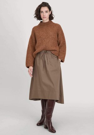 Femme aux cheveux bruns courts portant un pull brun épais, une jupe midi olive à taille à cordon et des bottes marron foncé arrivant au genou, se tenant sur un fond blanc.