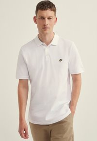 Camiseta polo blanca con un cuello acanalado, mangas cortas y un pequeño logo de un pájaro amarillo en el lado izquierdo del pecho. Textura de algodón suave. Diseño casual.