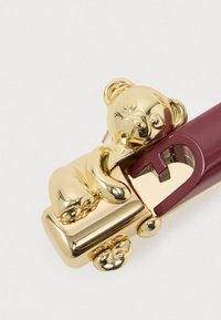 Aranyszínű medve figura, amely egy bordó és arany fém konzervnyitó végéhez van rögzítve, fehér háttéren.