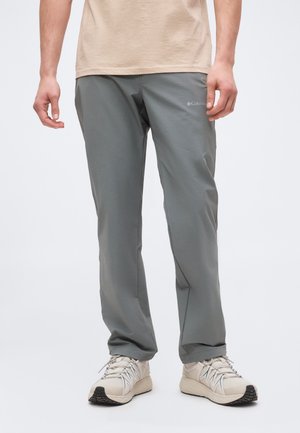 Homme portant un pantalon gris Columbia et des baskets beiges, debout devant un fond clair uni, mains détendues le long du corps.