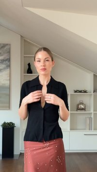 Camisa negra abotonada con mangas cortas y escote pronunciado; combinada con una falda coral con adornos blancos. Ambiente minimalista en la habitación.