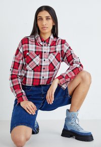 Chemise à carreaux rouge et noir à boutons avec deux poches poitrine, assortie à un short en denim bleu et des bottines bleu clair avec des semelles noires.