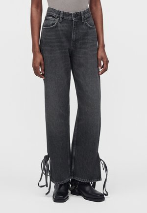 Person trägt hoch taillierte, weit geschnittene schwarze Denim-Jeans mit Bändern an den Säumen, kombiniert mit schwarzen Stiefeln mit quadratischer Spitze und einem grauen ärmellosen Oberteil.