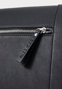 Sac en cuir noir doté d'une fermeture éclair métallique avec un tirette en argent. Comprend une surface texturée et une étiquette de fermeture éclair estampillée.