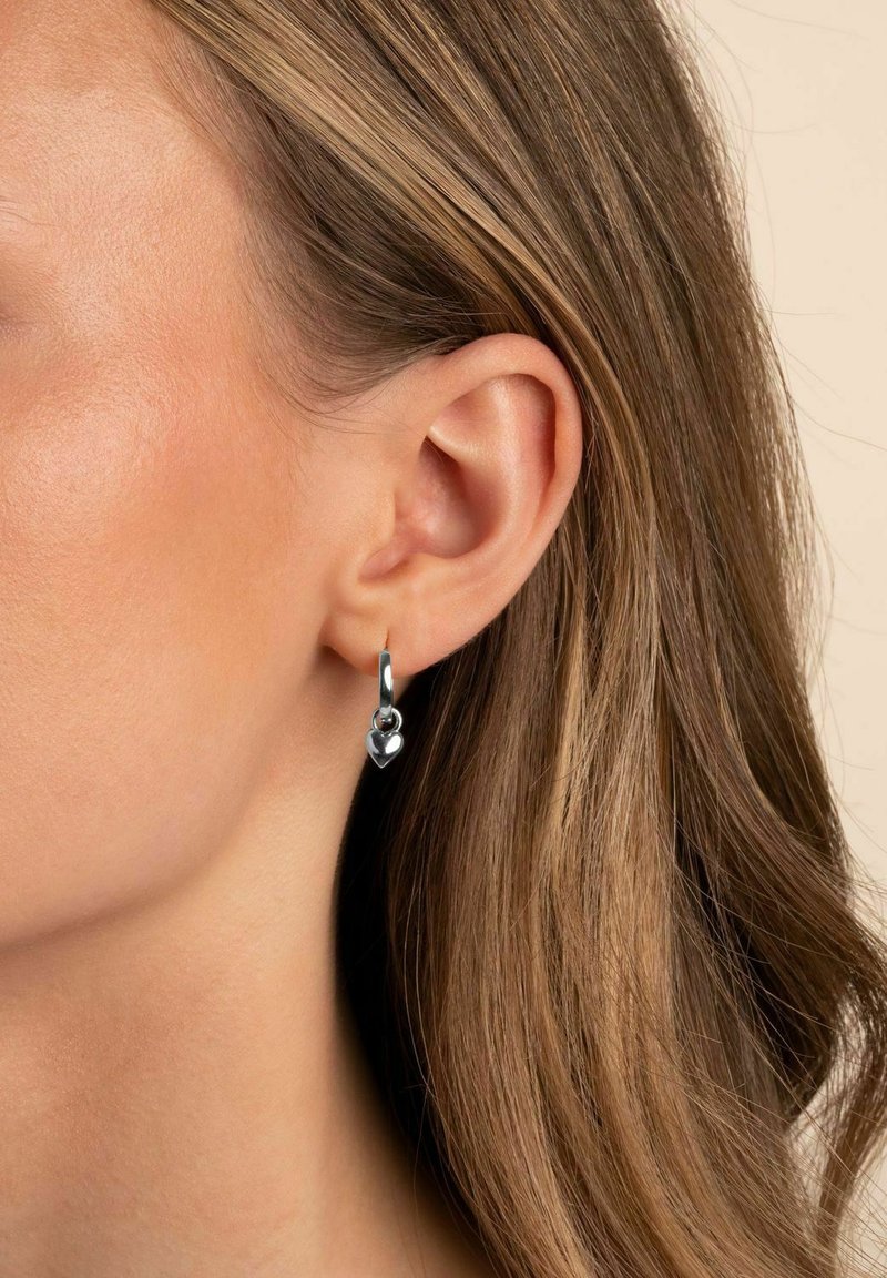 Silverörhänge med en hjärtanhänge, med en polerad yta och en enkel låsning, bärs på en modells öra.