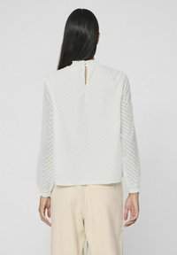 Blusa bianca a maniche lunghe con motivo a chevron testurizzato, scollatura rotonda, dettaglio apertura a chiave sul retro e polsini elasticizzati. Abbinata a pantaloni beige chiaro.