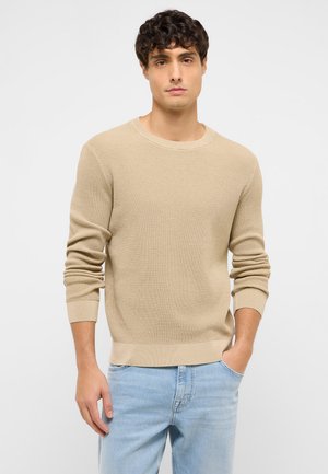 Mann mit kurzen lockigen Haaren, der einen beigen Strickpullover und hellblaue Jeans trägt, steht mit einer Hand in der Tasche vor einem schlichten Hintergrund.