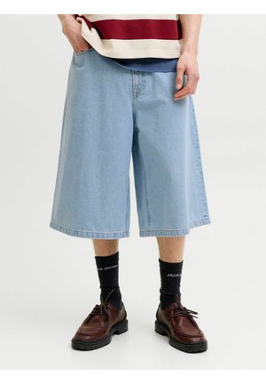 Personne portant un short en jean bleu clair à jambes larges arrivant au genou, des chaussettes noires avec texte blanc, et des chaussures marron en cuir à lacets.