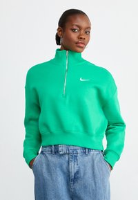 Sweatshirt verde com fecho, gola alta e punhos elásticos, apresentando um pequeno logo branco da Nike no peito, combinado com calças de ganga azuis.