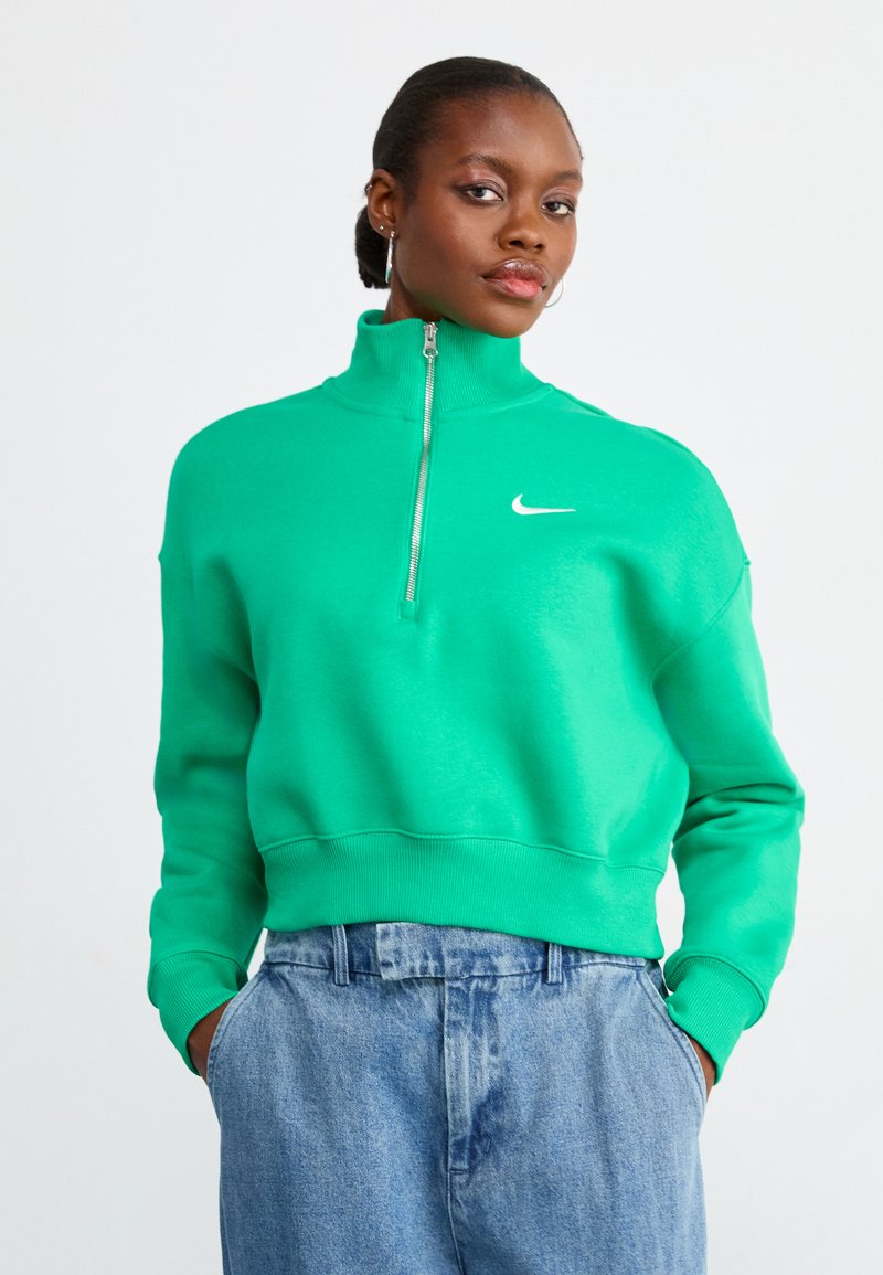 Sweatshirt verde com fecho, gola alta e punhos elásticos, apresentando um pequeno logo branco da Nike no peito, combinado com calças de ganga azuis.