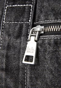 Tecido de denim preto com um fecho éclair prateado. O fecho tem uma aba de puxar retangular com "lala BERLIN" gravado, com detalhes em costura branca.
