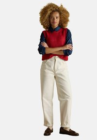 Woman with curly hair stands with arms crossed wearing red sleeveless sweater, navy shirt, cream pants, and brown loafers on white background.
