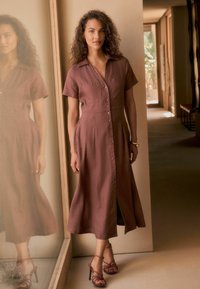 Gaun coklat dengan lengan pendek dan kancing, dengan bodice yang ramping dan rok yang melebar. Dipadukan dengan sandal hak tinggi yang bersarung. Latar belakang cermin reflektif.