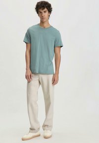 Lichtblauwe katoen t-shirt met korte mouwen, gecombineerd met crèmekleurige recht-toe-recht-aan broek en off-white sneakers, tegen een neutrale achtergrond.