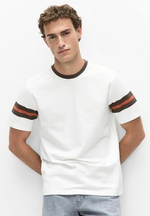Jeune homme aux cheveux courts et bouclés pose devant un fond uni, vêtu d’un t-shirt blanc avec des rayures marron et orange sur les manches et d’un jean bleu.
