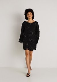 Melnas kleitas ar garām zvana piedurknēm, caurspīdīga ziedu raksts un taisns siluets, valkāta ar melniem plakanajiem sandaļiem. Gluda tekstūra, līdz viduklim garums.