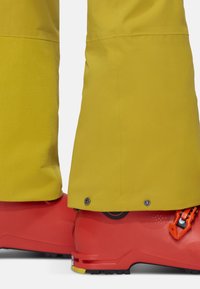 Gele waterdichte skibroek heeft een textuur in het onderste gedeelte, drukknoopsluitingen en wordt gecombineerd met felrode skischolen met oranje accenten.