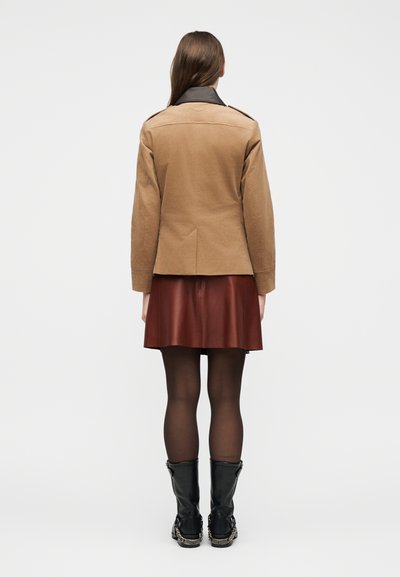 Γυναίκα που στέκεται με το βλέμμα προς τα πίσω, φορώντας ένα μπεζ σακάκι με σκούρο γιακά, καφέ δερμάτινη φούστα, διαφανή μαύρα καλσόν και μαύρες μπότες μέχρι τη μέση της γάμπας με λεπτομέρεια αλυσίδας.
