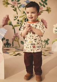 Kortærmet skjorte med et legende dyreprint i flere farver, kombineret med brune cordbukser og tan-farvede støvler. Baggrunden har blomstermønstre.