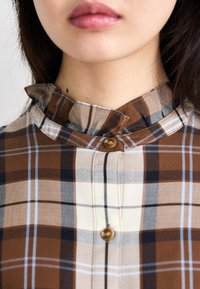 Bruine geruite blouse met knopen, voorzien van een ruches-kraag, gouden knopen en een textiel met afwisselend lichte en donkere vierkanten.