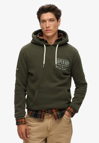 Olivengroene katoenen hoodie met verstelbare trekkoorden, voorzak en logo. Gelaagd over een zwart en oranje geruite shirt, gedragen met beige broek.