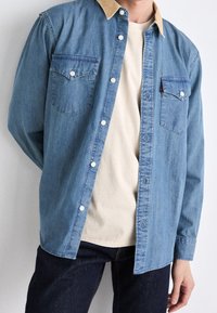 Camicia di jeans azzurro chiaro con colletto in corda beige, due tasche sul petto e bottoni argentati, indossata sopra una t-shirt beige e jeans scuri.