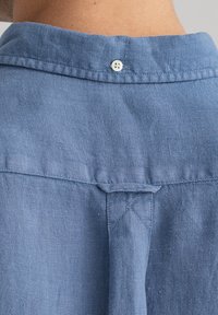 Camicia in lino azzurro chiaro con colletto a punta, dotata di un singolo bottone bianco sul retro, cuciture visibili e una finitura textured.