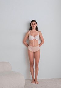 Lichtroze kanten beha en naadloze beige hoog opgesneden slip op een model, staand tegen een lichtgrijze muur, met een zacht textuur vloerkleed in de buurt.