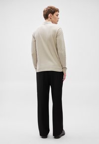 Bež dolg rokav polo majica z mehko teksturo, kombinirana z ohlapnimi črnimi hlačami. Preprost dizajn, prikazan od zadaj na nevtralnem ozadju.