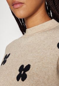 Beiger Strickpullover mit hohem Kragen und schwarzen Blumenmustern. Die Textur wirkt weich und gemütlich und betont das Strickmaterial.