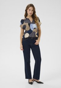 Kratka bluzica s kratkimi rokavi z večbarvnim abstraktnim vzorcem v modrih, rjavih in bež tonih; v kombinaciji s temno modrimi ravnimi džinsami in črnimi petkami.