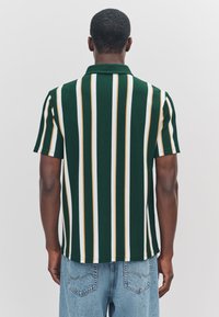 Grünes, vertikal gestreiftes Poloshirt mit gelben und weißen Akzenten; gerippte Textur. Kombiniert mit hellblauen Jeans aus Denim.